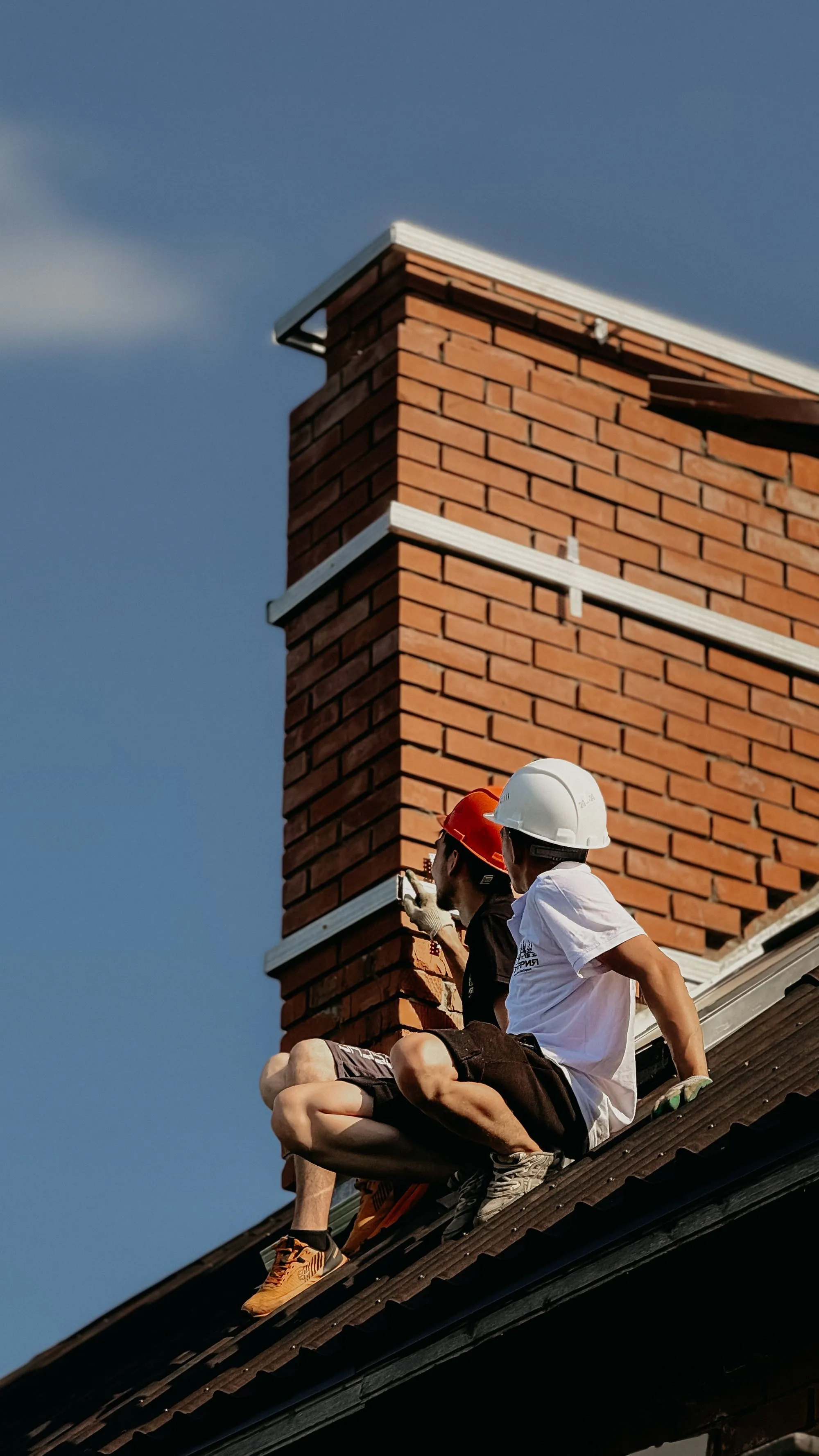 Two workers in hard hats on a roof beside a tall brick chimney