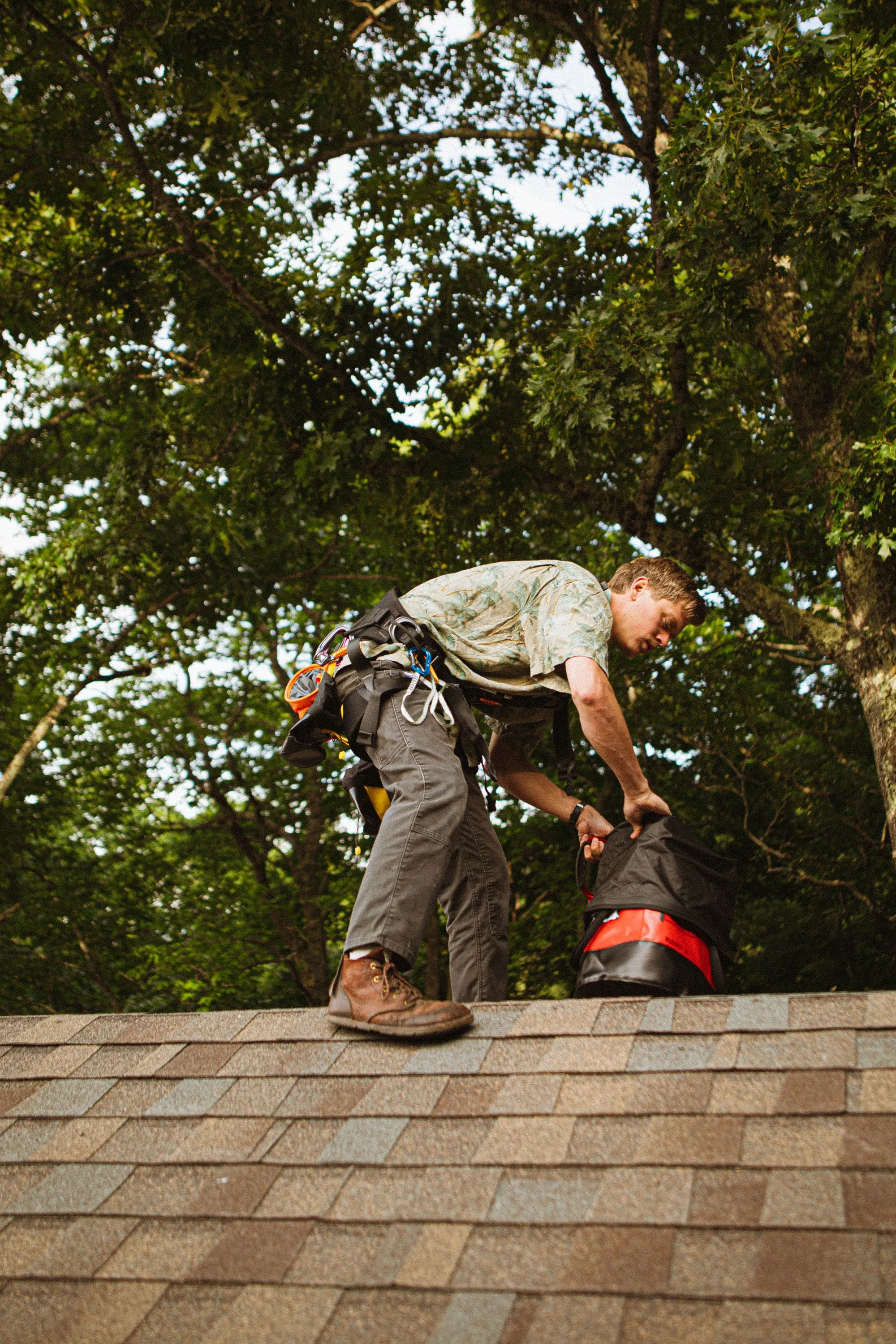 A roofer in a harness loading debris into a bag on a stripped roof