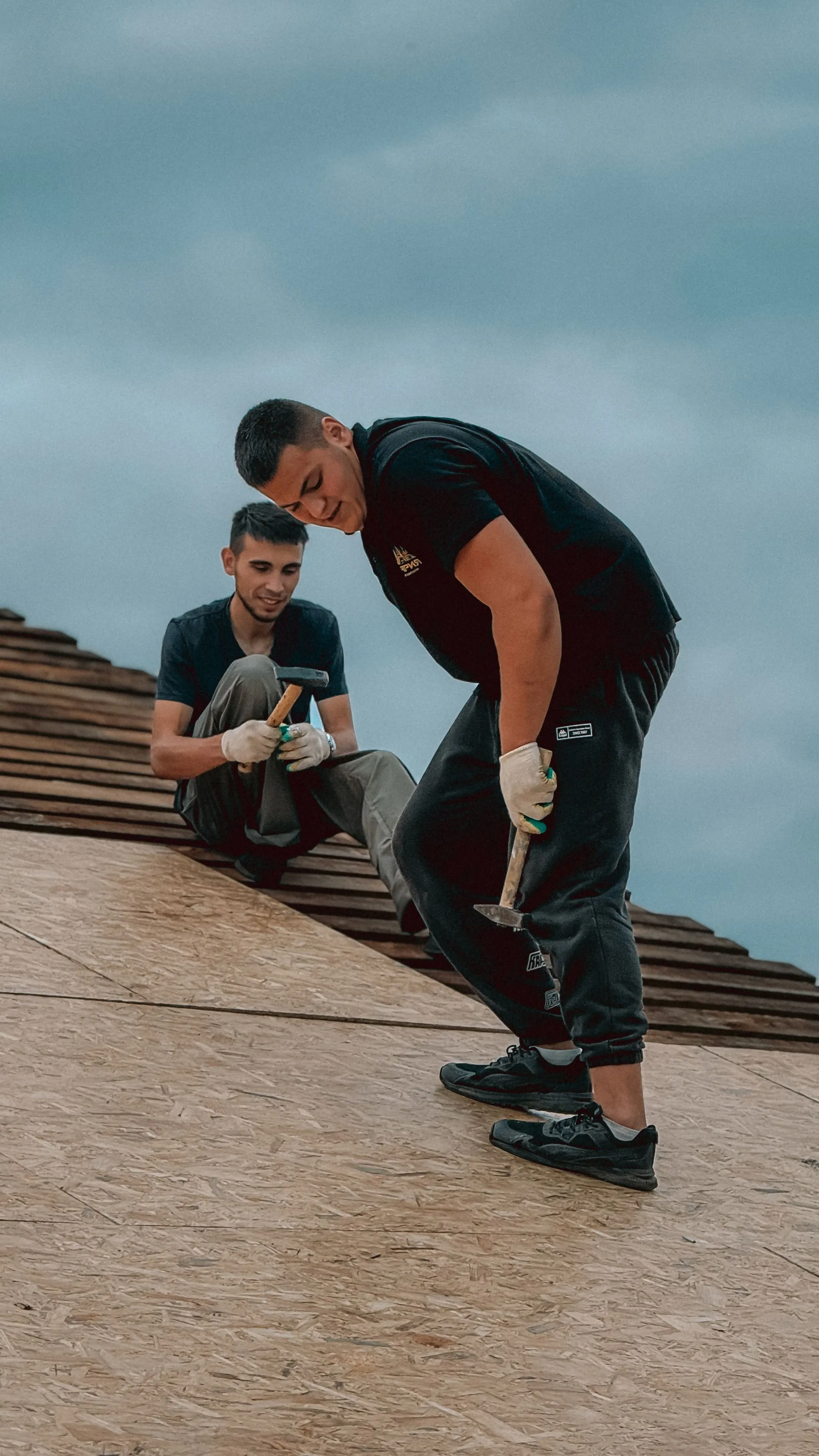 A roofer kneeling on bare roof sheathing, driving a nail by hand