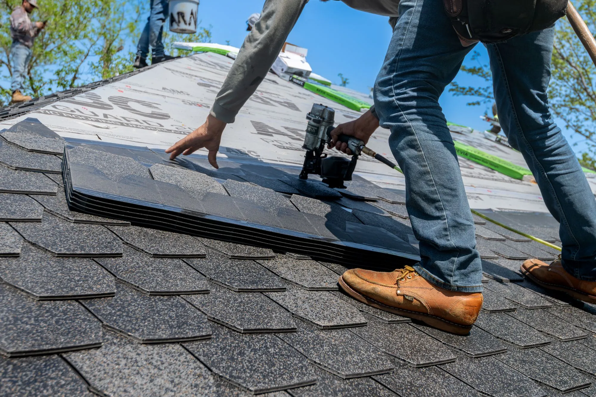 A roofer running a nail gun along a fresh course of asphalt shingles