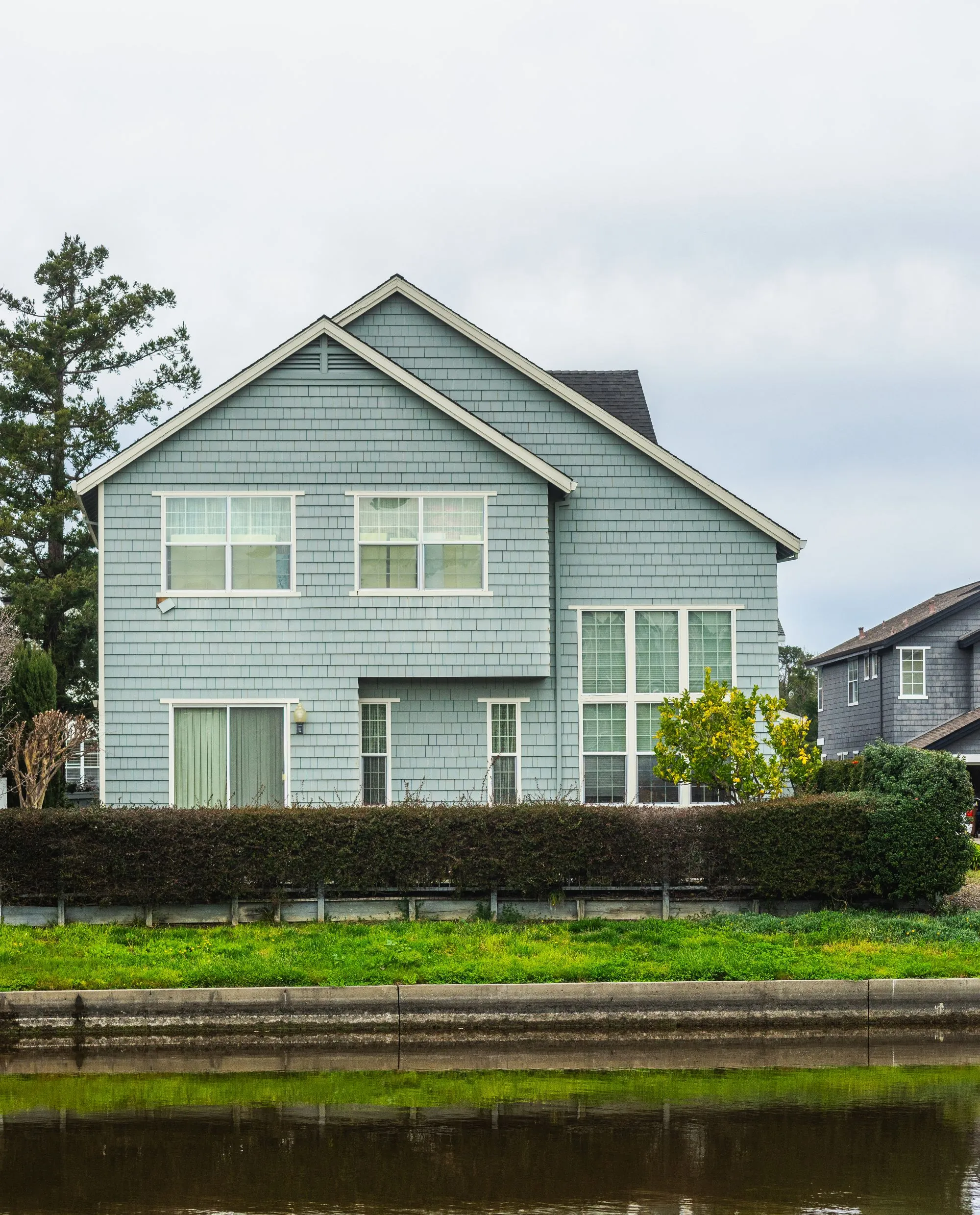 A pale blue two-storey house with a shingled roof and a hedge along the front
