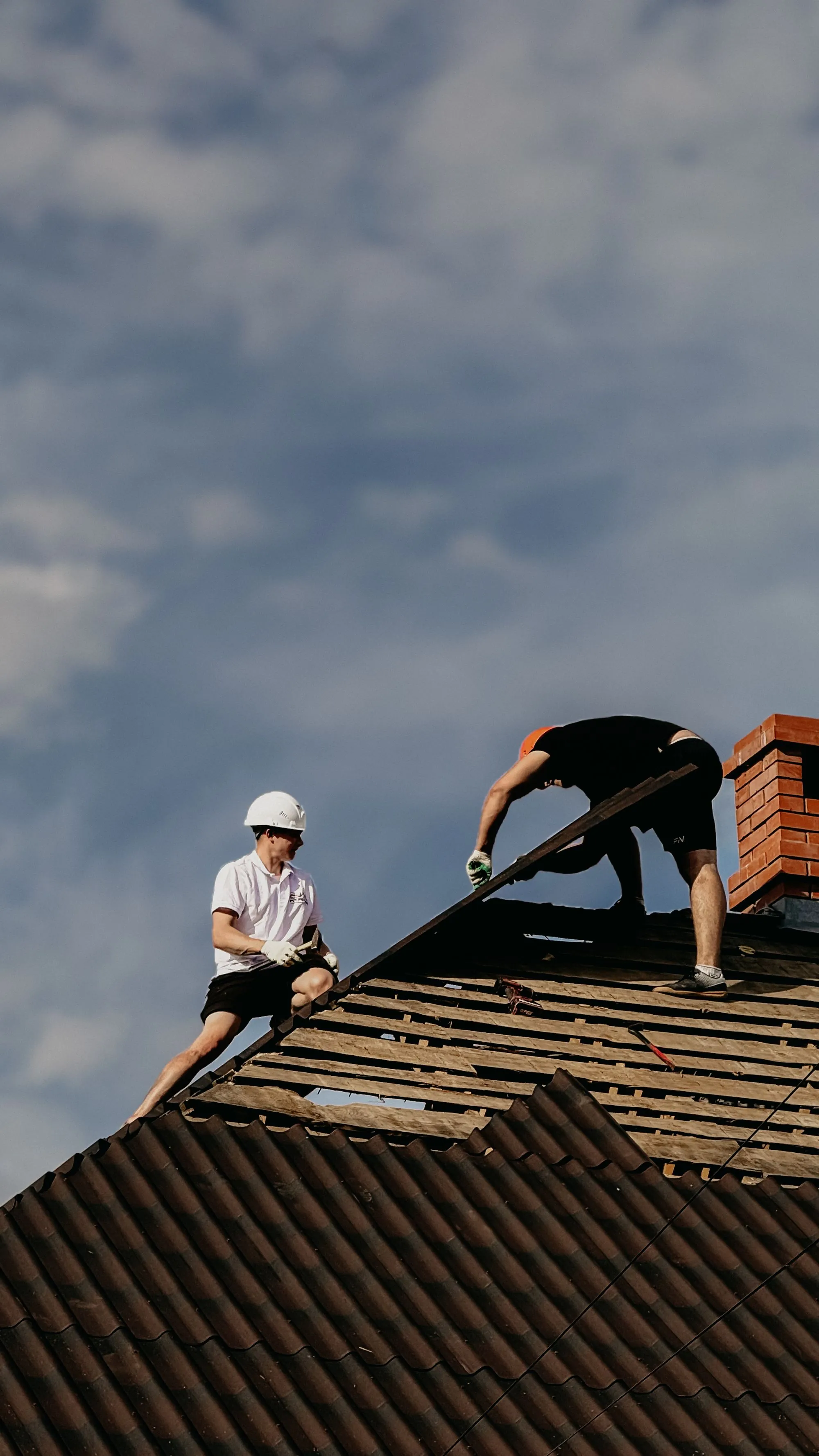 Two roofers working their way along a dark roof slope beside a brick chimney